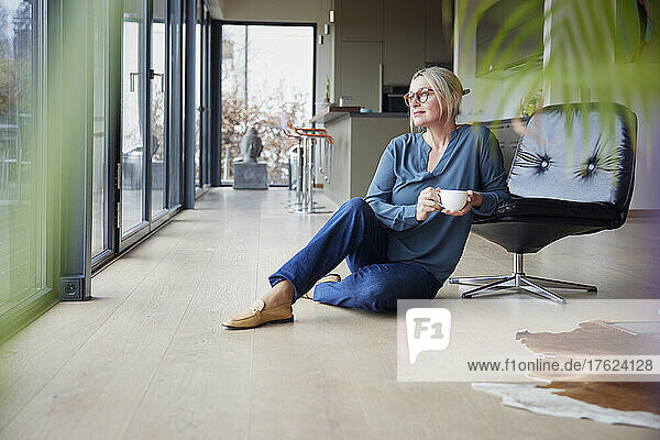 Blond woman with coffee cup sitting by chair at home