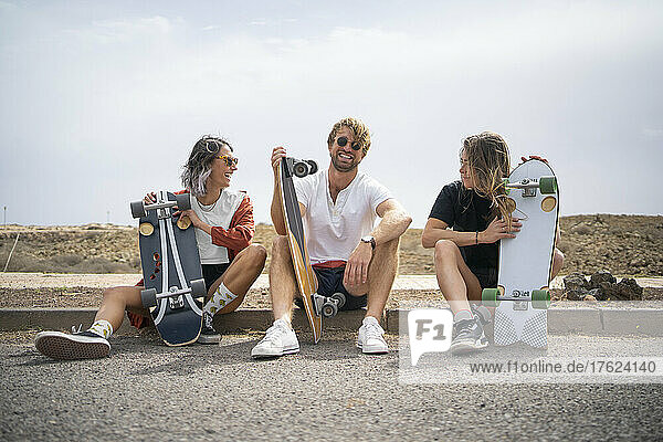 Happy friends with skateboards sitting at roadside on sunny day