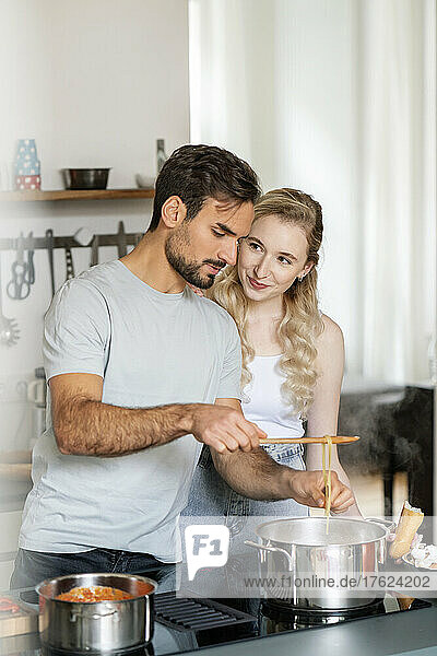 Smiling blond woman looking at boyfriend making pasta in kitchen