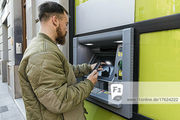 Junger Mann mit Smartphone benutzt Geldautomaten