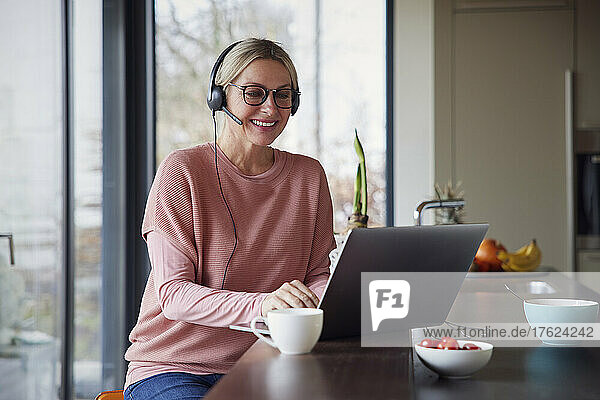 Happy blond woman wearing headphones using laptop in kitchen