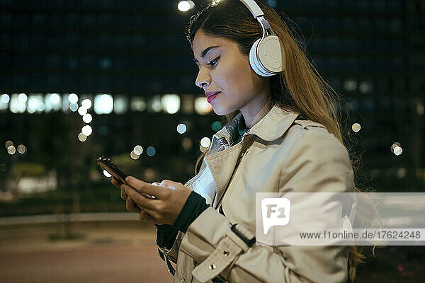 Beautiful woman using phone listening music through wireless headphones at night