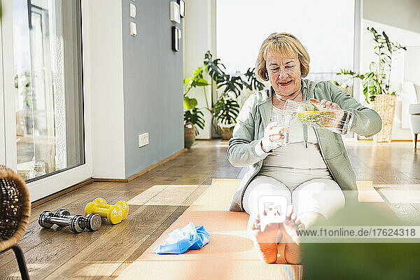Blond senior woman pouring water in glass sitting on exercise mat at home