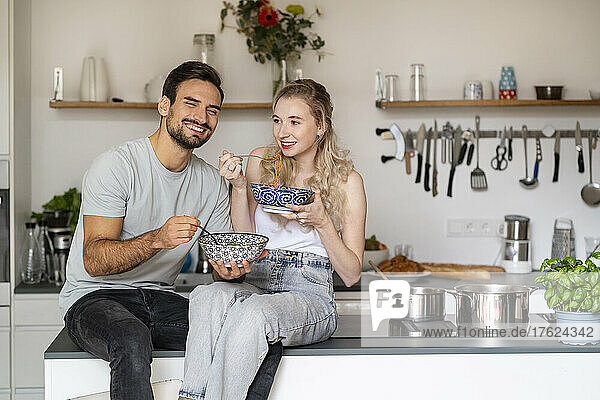 Smiling young woman and man eating spaghetti sitting on kitchen island at home