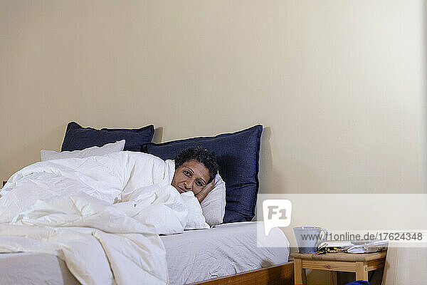 Woman lying on bed covered in white blanket at home