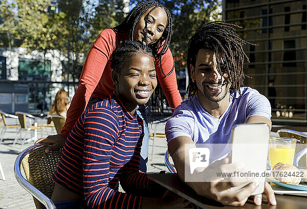 Happy young man with friends taking selfie through mobile phone at sidewalk cafe