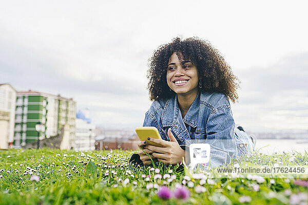 Lächelnde Frau mit Smartphone träumt auf der Wiese