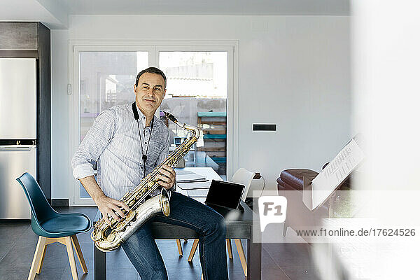 Smiling musician with saxophone sitting on table at home