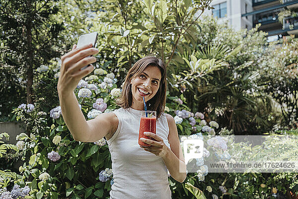 Lächelnde Frau macht Selfie mit Smartphone und Smoothie-Glas im Garten