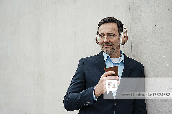 Businessman holding disposable coffee cup listening music through wireless headphones by wall