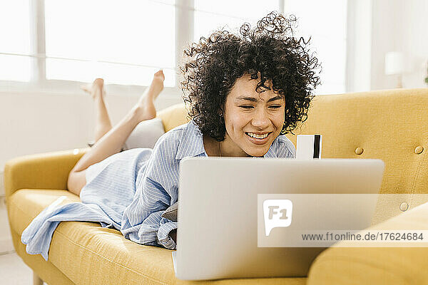 Smiling young woman with laptop and credit card lying on sofa at home
