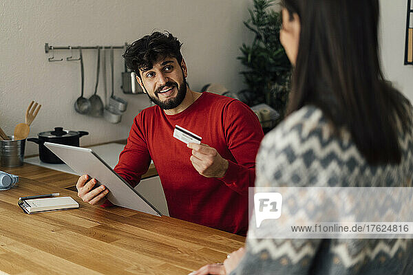 Man holding tablet PC showing credit card to girlfriend in kitchen at home