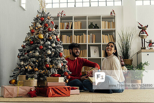 Happy woman with boyfriend holding gift box sitting by Christmas tree at home
