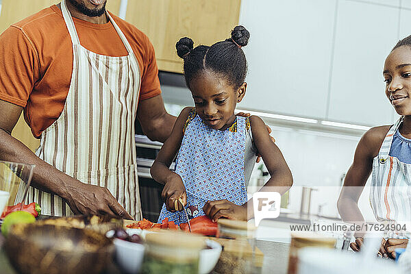 Girl cutting pointed pepper by father and sister in kitchen