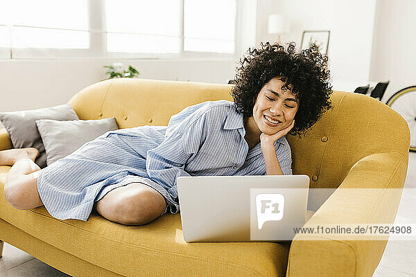 Lächelnde junge Frau mit Laptop  die zu Hause auf dem Sofa im Wohnzimmer liegt