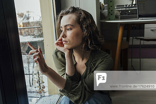 Sad woman with head in hand sitting by glass window at home