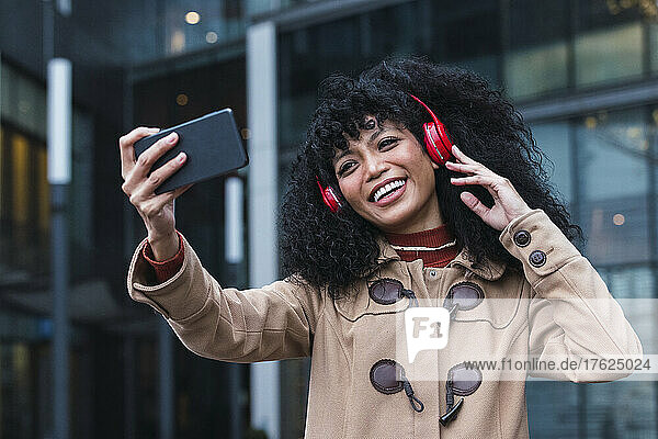 Cheerful woman taking listening music through wireless headphones taking selfie through smart phone in city