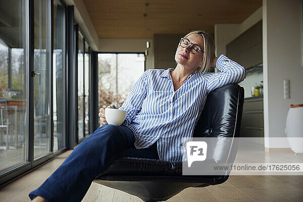 Blond woman with eyes closed holding coffee cup sitting on chair at home