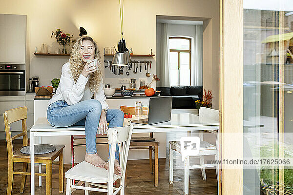 Cheerful blond woman holding coffee cup sitting on table in kitchen