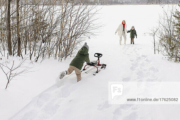 Woman with son looking at boy pushing toboggan on snow in winter
