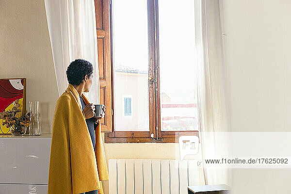 Woman wrapped in blanket holding mug and looking out of window at home