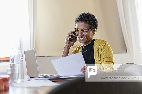 Smiling freelancer with paper document talking on smart phone at home