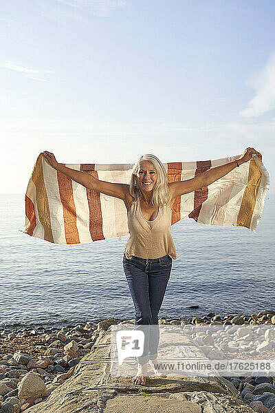 Happy woman with arms outstretched holding blanket enjoying at beach on sunny day