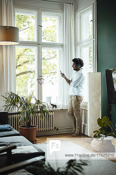 Happy man using mobile phone and listening music through wireless headphones by window at home