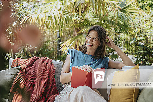 Glückliche Frau mit Buch sitzt auf Sofa im Hinterhof