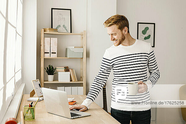 Glücklicher junger Mann mit Kaffeetasse und Laptop  der am Tisch im Wohnzimmer steht