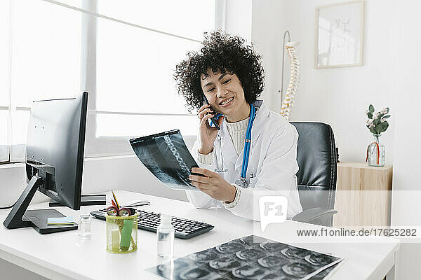 Smiling young doctor holding X-ray talking on mobile phone sitting at desk in hospital