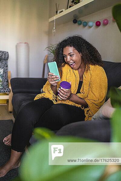 Happy young woman with mouth open using smart phone on sofa in living room at home