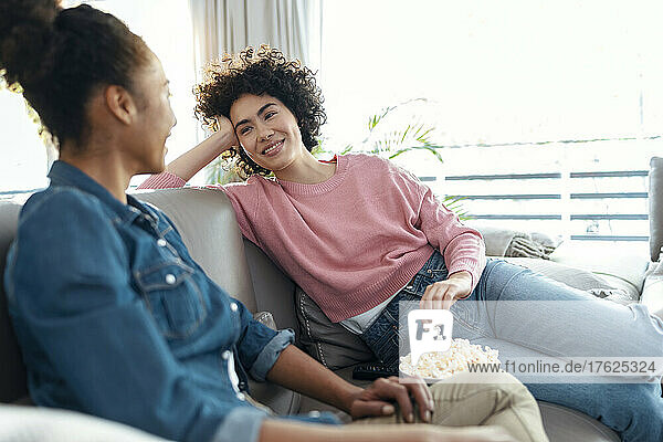 Smiling young woman with popcorn looking at friend sitting on sofa in living room