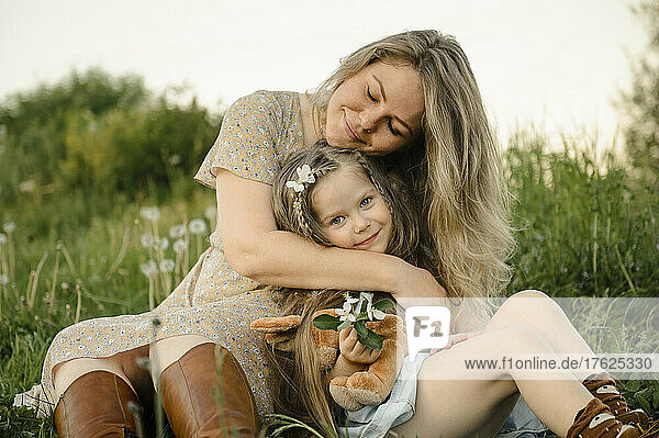 Smiling mother with eyes closed embracing cute daughter sitting in meadow