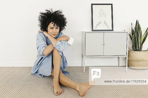 Smiling young woman with coffee cup sitting on ground by white wall at home