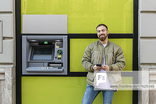 Lächelnder junger Mann mit Smartphone am Geldautomaten