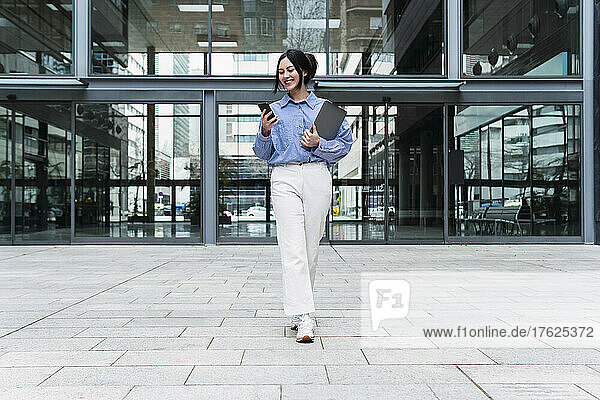 Smiling businesswoman holding laptop using mobile phone walking in front of office building