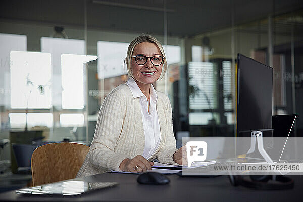 Smiling businesswoman wearing eyeglasses sitting at desk in office