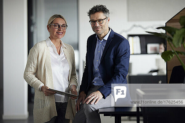Smiling businesswoman with tablet PC standing by colleague sitting on desk in office