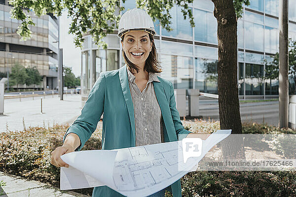 Happy architect wearing hardhat standing with construction plan