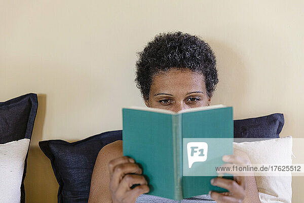 Woman with black short hair reading book on bed at home