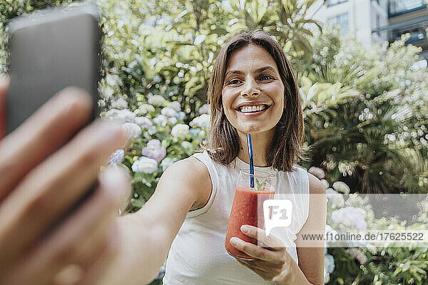 Happy woman taking selfie holding smoothie glass in garden