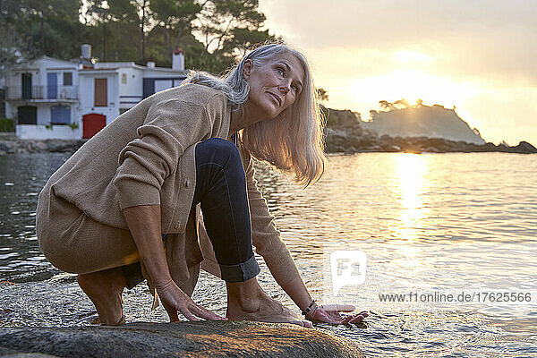 Gray haired woman dipping hand in water on rock at beach