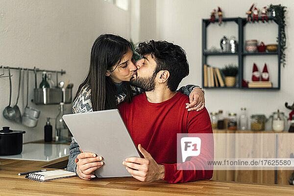 Mann hält Tablet-PC und küsst Freundin zu Hause in der Küche