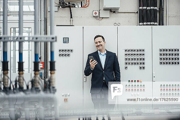 Happy businessman with hand in pocket using smart phone in front of machinery