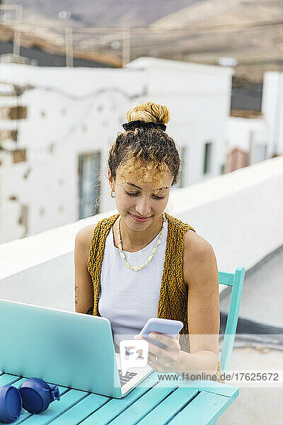 Smiling woman surfing net through mobile phone sitting with laptop at table on rooftop