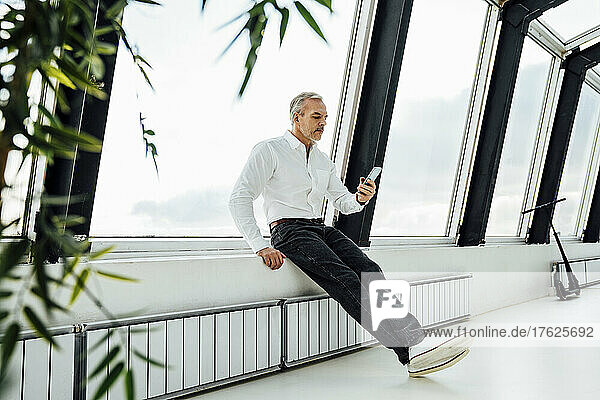 Businessman using smart phone sitting on window sill at work place