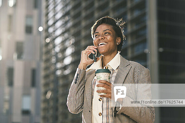 Cheerful businesswoman holding coffee cup talking through mobile phone on sunny day