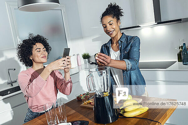 Young woman photographing friend making juice in kitchen at home