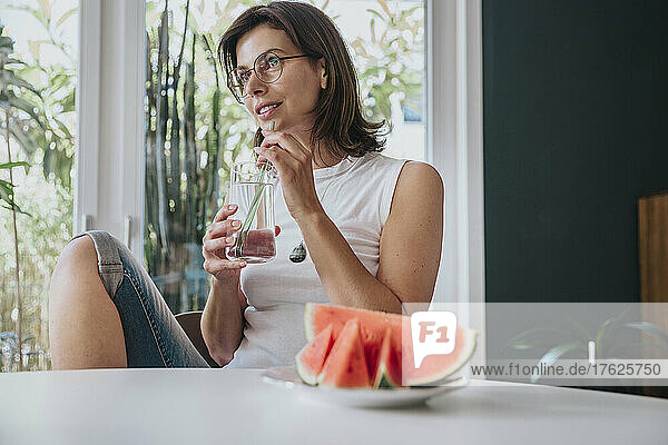 Thoughtful woman drinking water with watermelon on table at home